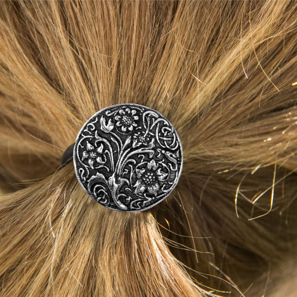 Round silver ponytail holder with a detailed wildflower and vine embossed design, secured in blonde hair.